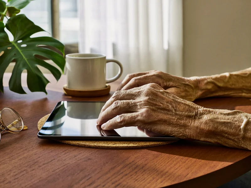 Altersgezeichnete Hände ruhen auf einem Tablet neben Keramiktasse, Pflanze und Lesebrille.